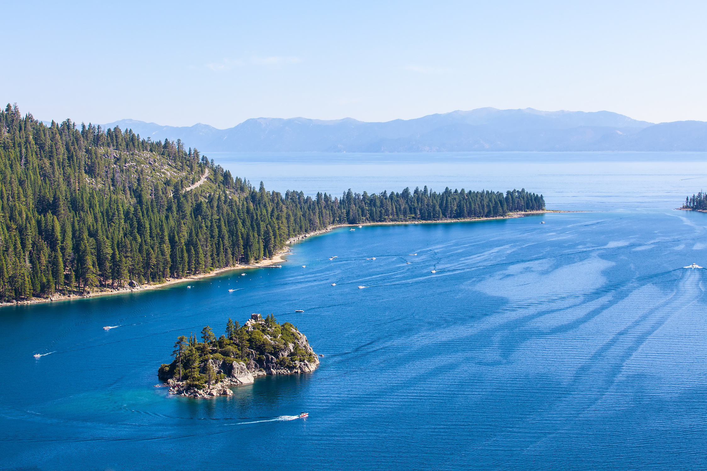 An aerial view of Lake Tahoe with blue water and evergreen trees growing along a hill. There's in island in the middle of the bay and a few boats in the water.